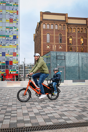 Un père se déplace en zone urbaine avec son fils sur un vélo cargo shortail.