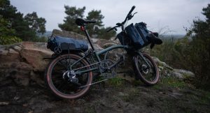 Un vélo pliant Brompton G-Line équipé de sacoches de voyage appuyé contre un rocher dans un décor forestier.