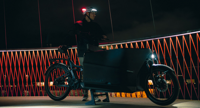 Un homme se tient à côté d'un biporteur Riese&Müller coiffé d'un casque Abus Hyp-E feux allumés sur un pont urbain en pleine nuit.