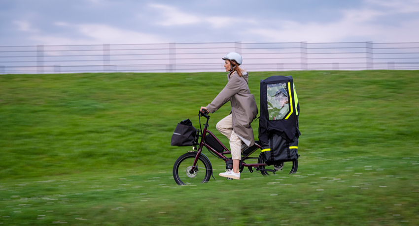 Une femme au guidon d'un longtail Tern traverse un paysage d'herbe humide. A l'arrière, un enfant est installé sur le porte-bagages sous une tente de pluie.